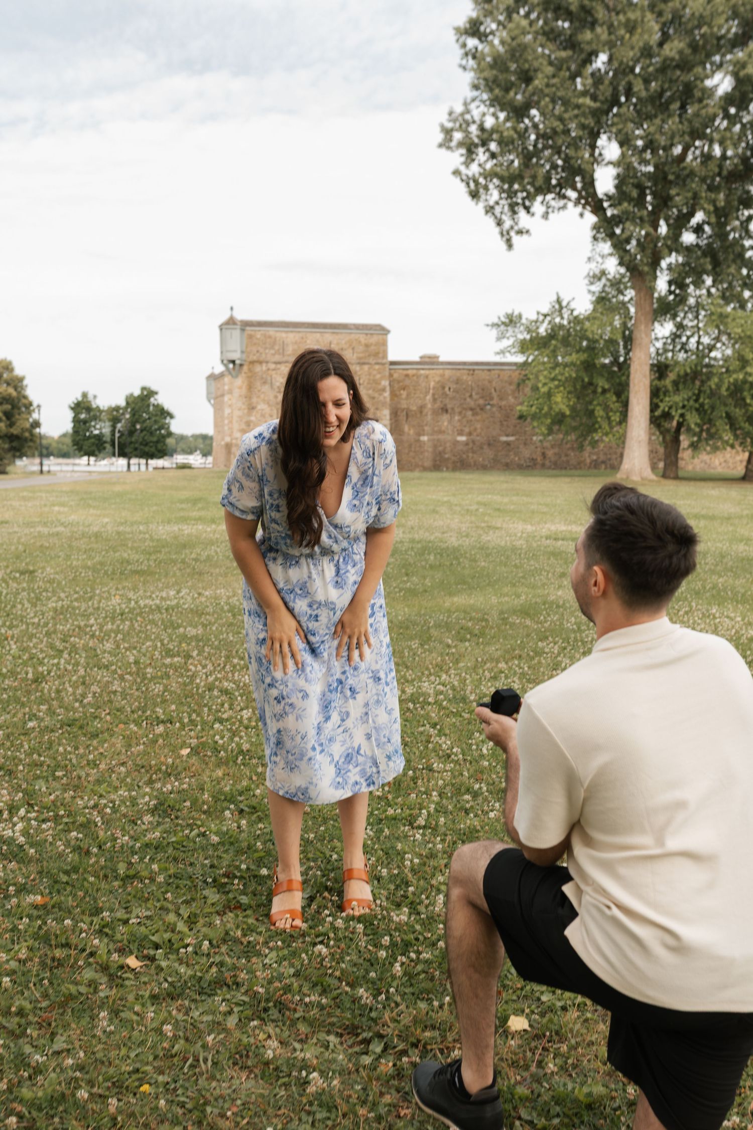 Man proposing to woman in blue floral dress outdoors on green grass near historic brick building.