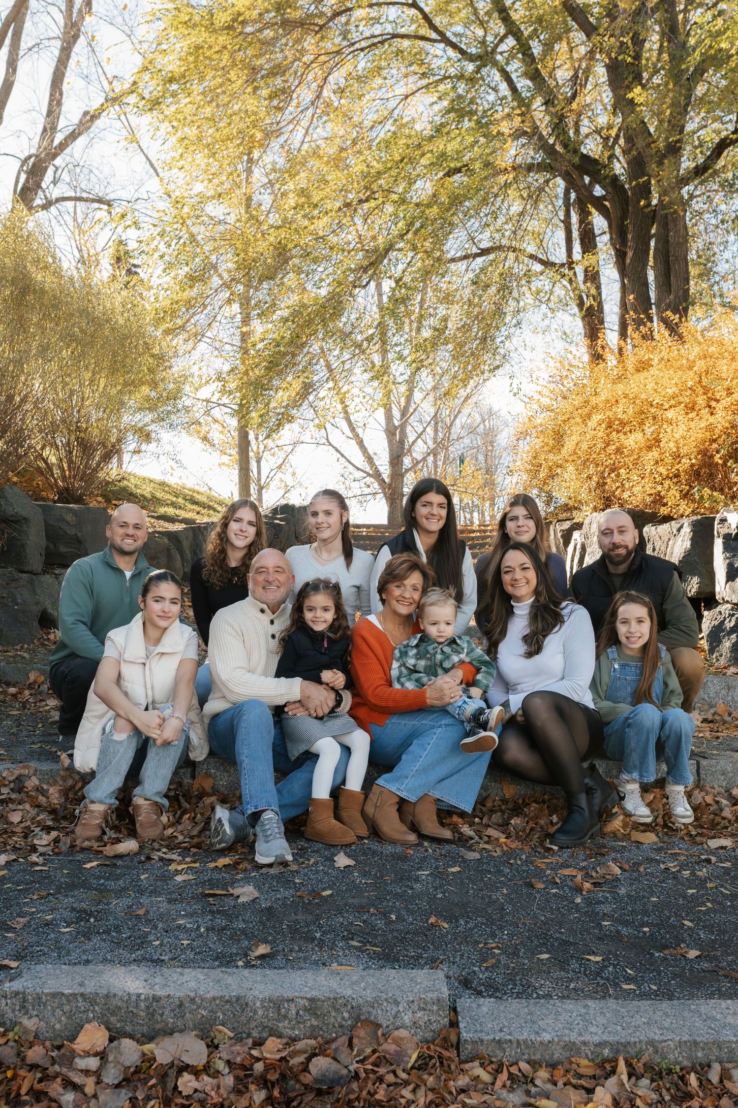 Large multigenerational family posing together outdoors in autumn, surrounded by colorful fall foliage and golden trees.
