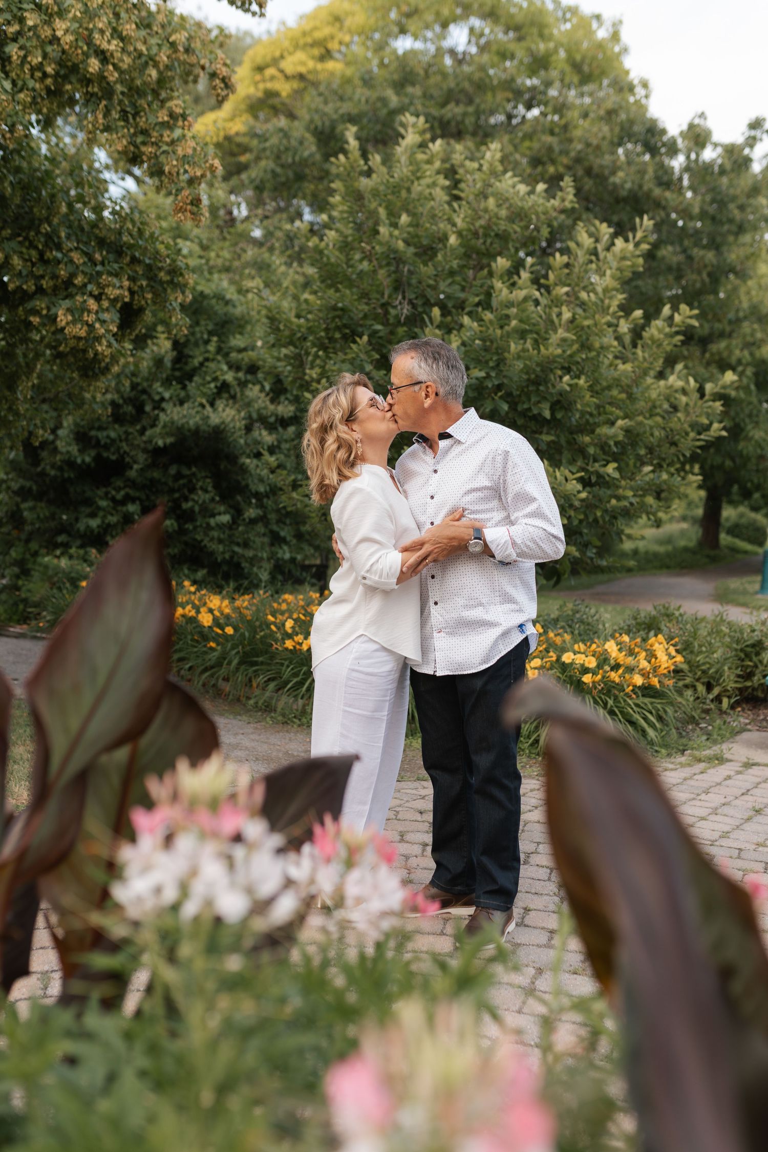 Mature couple sharing a kiss in a lush garden surrounded by colorful flowers and green trees.