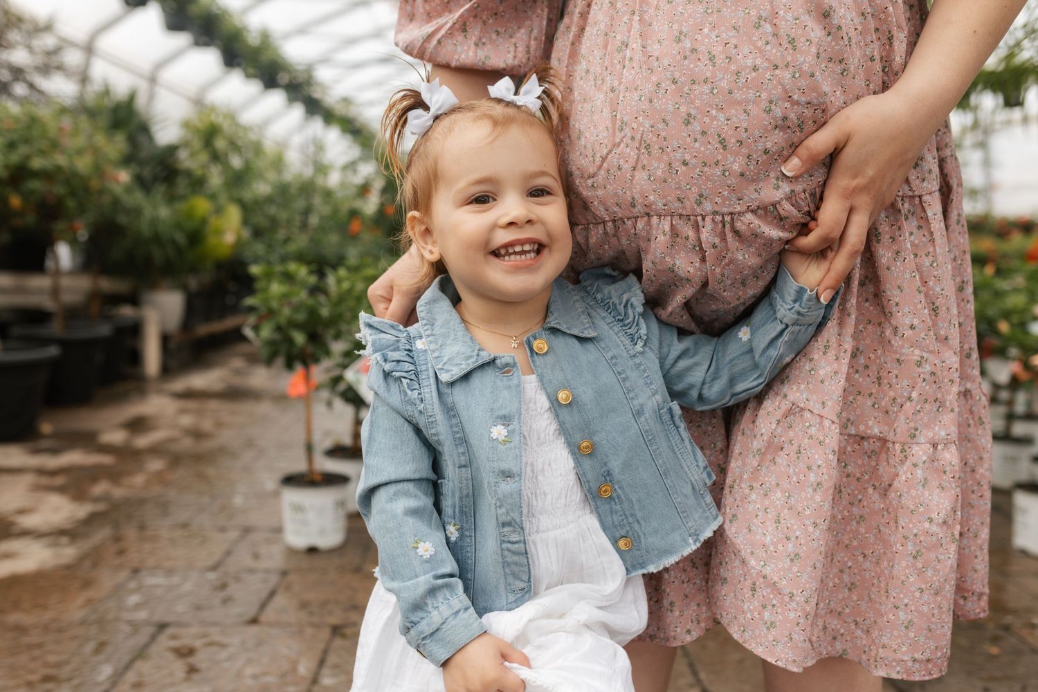 A laughing toddler girl in a denim jacket stands beside a pregnant woman in a floral dress inside a greenhouse.