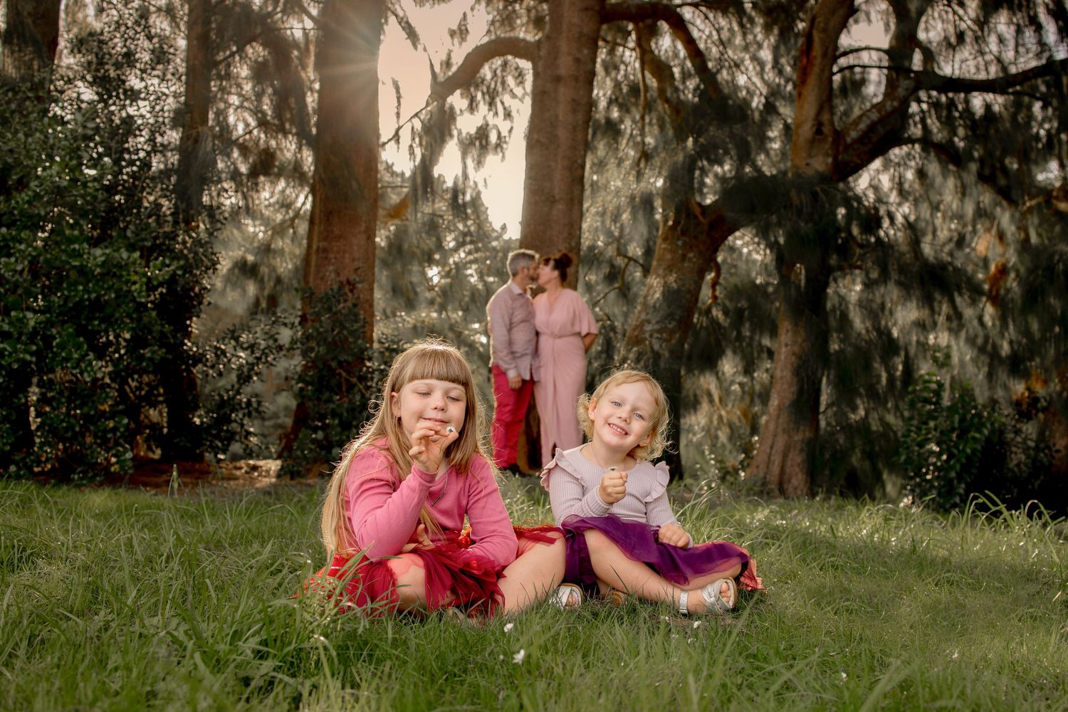Young sisters sitting on grass in foreground, parents embracing in background, sun shining through the trees. Outdoor family photoshoot.