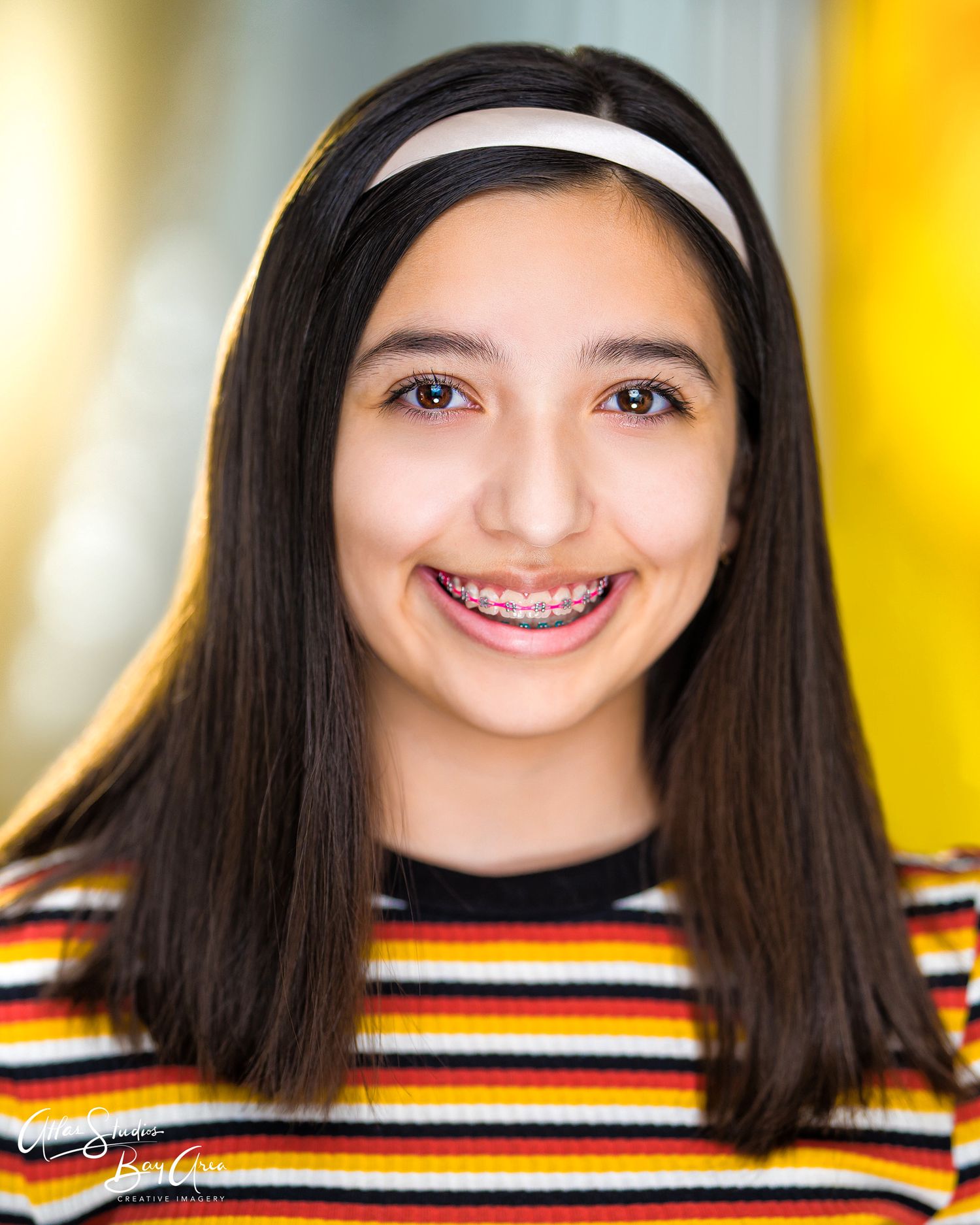 A smiling individual wearing braces and a striped shirt poses for a headshot against a blurred yellow background.