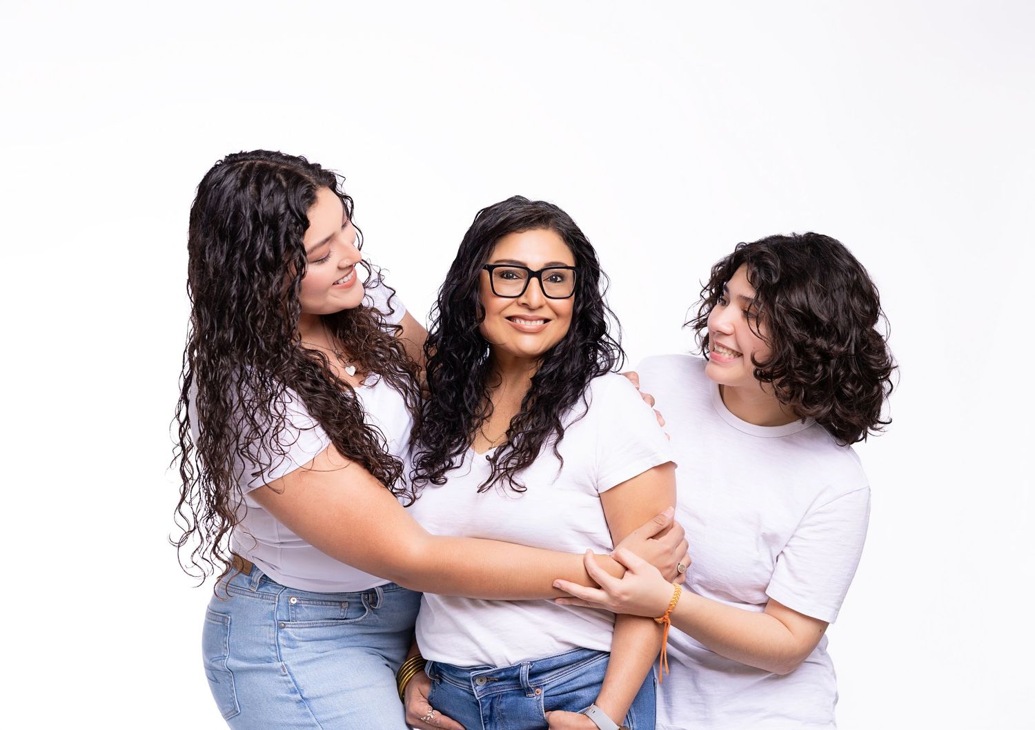 Three people dressed casually in jeans and white tops stand together smiling and embracing against a white background.
