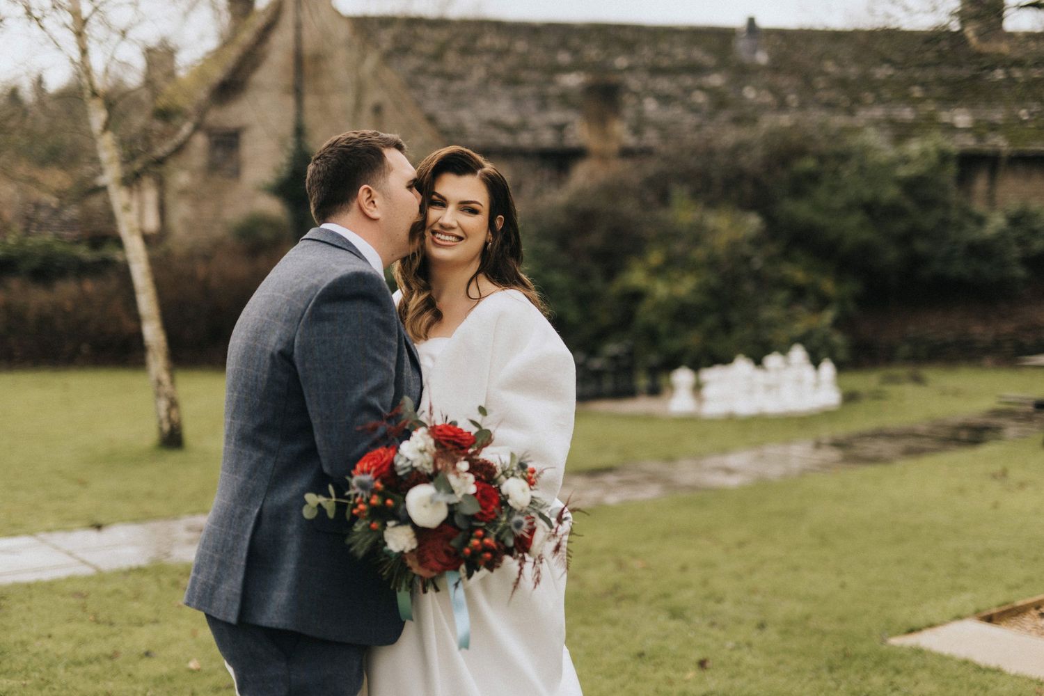 A couple shares an intimate moment outdoors on their wedding day, with the bride holding a vibrant red bouquet.