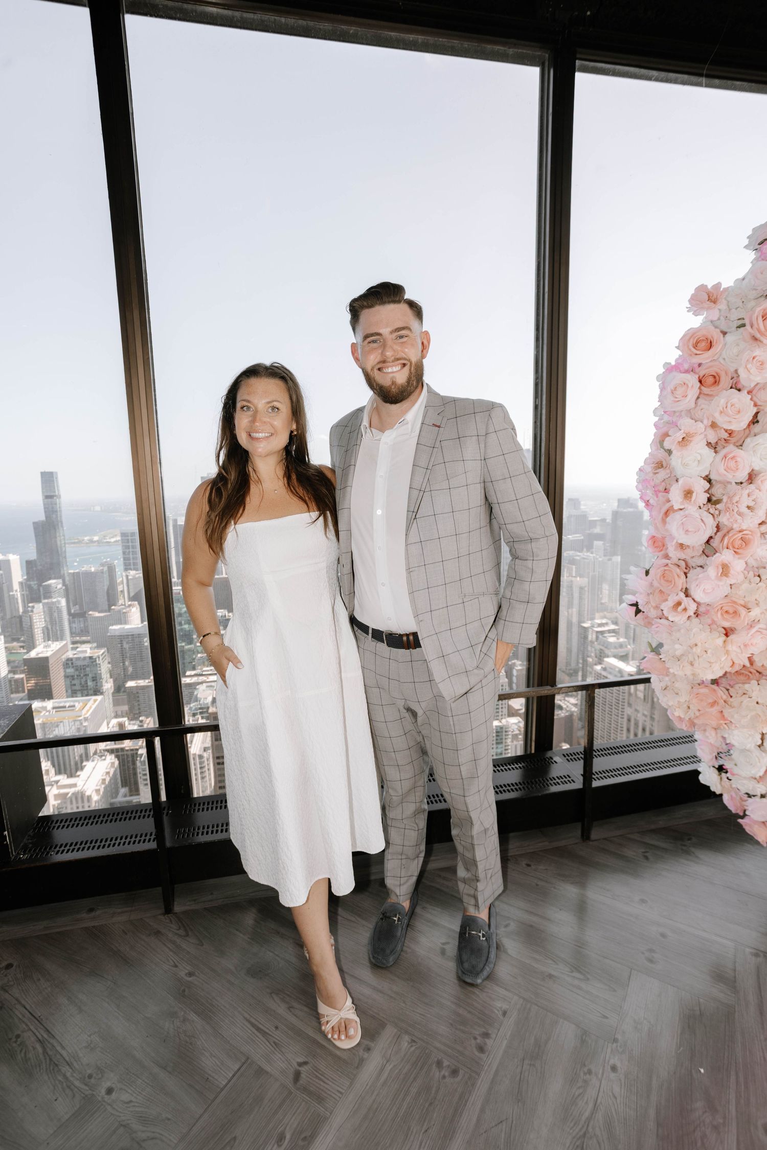 A couple poses together against floor-to-ceiling windows with a city skyline view and pink floral decorations.