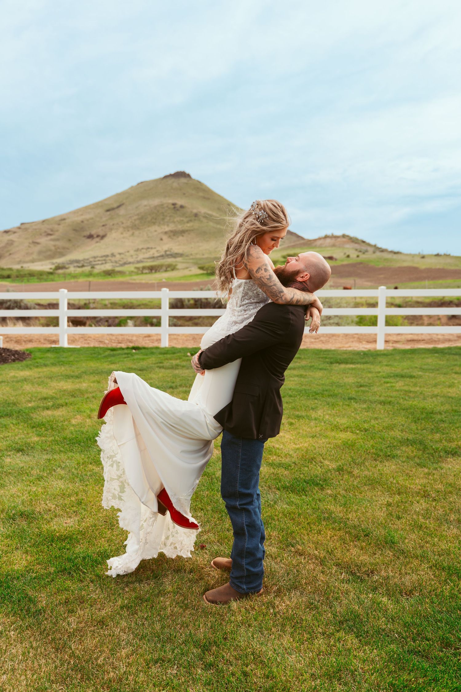 A couple shares a romantic dip pose in front of rolling hills and a white fence on a green grassy field.