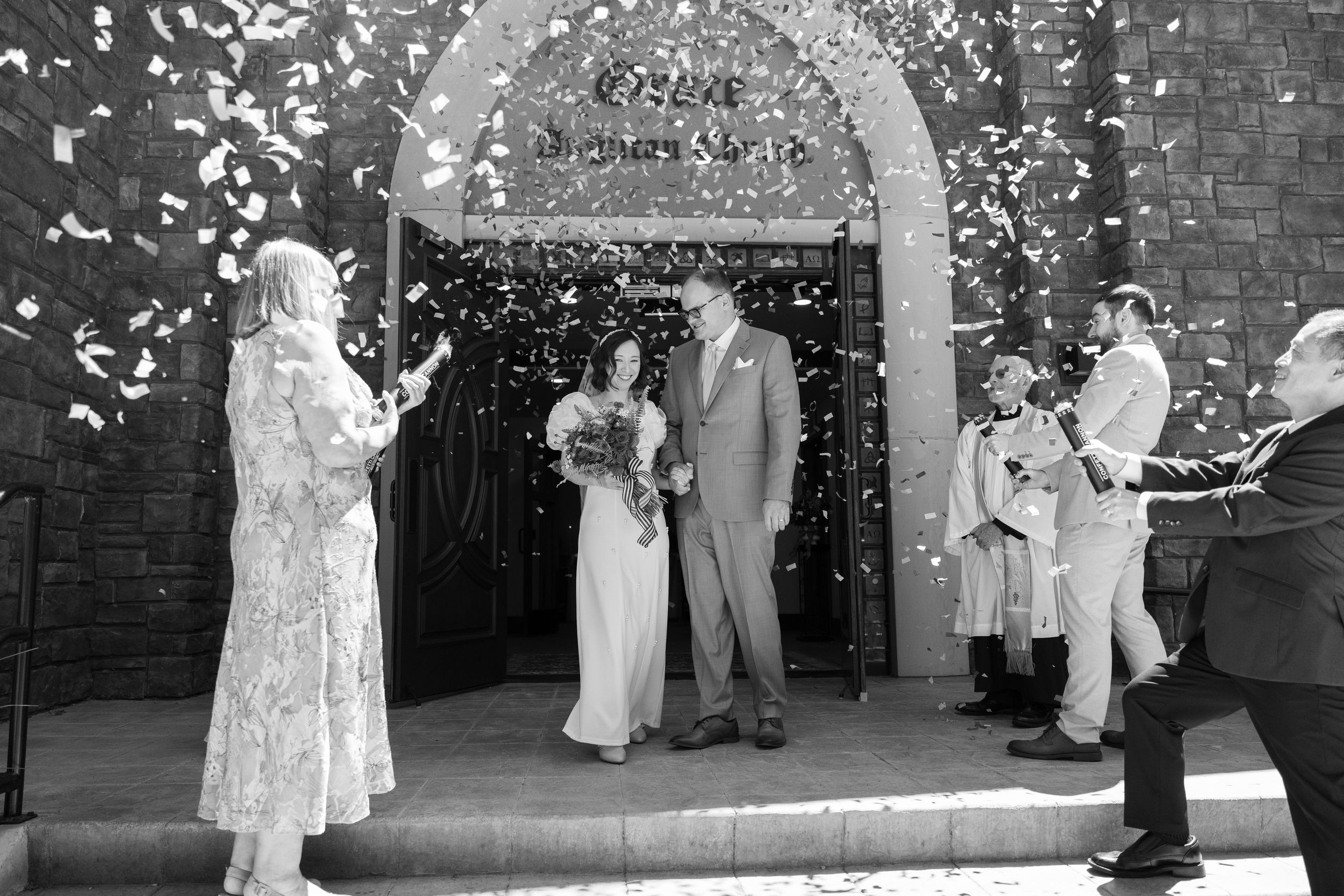 A wedding ceremony under a stone archway with confetti falling as guests look on.