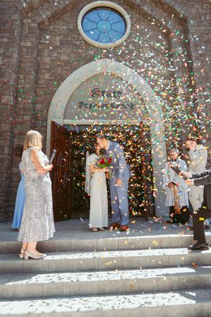 Wedding celebration outside a stone church with confetti flying as newlyweds exit through an arched doorway on stone steps.