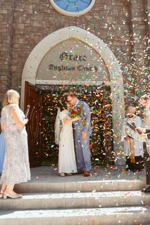 Couple stands under brick church archway while colorful confetti rains down during wedding celebration.
