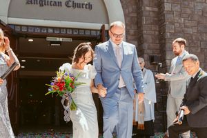 Wedding party celebrates joyfully outside a brick Anglican church with raised arms and flowers.