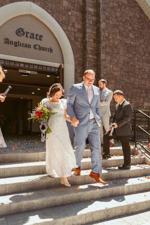 Newlyweds exit Grace Anglican Church down stone steps while guests shower them with confetti in the bright sunlight.