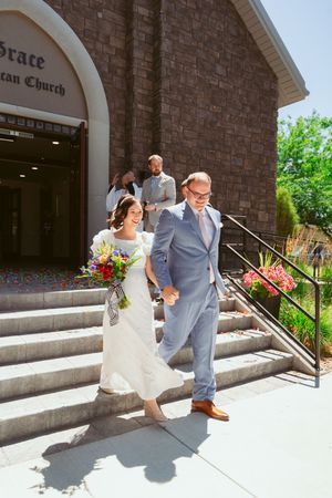 A couple exits a brick church on their wedding day, descending stone steps together in the bright sunlight.