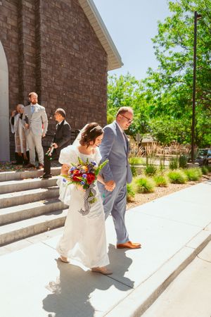 Wedding party descends church steps on sunny day with bride carrying vibrant bouquet and groom in light gray suit.