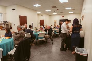 People gather in a large community room with teal tablecloths and tables set up for a banquet-style event.