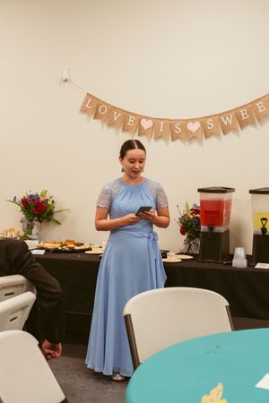 Guest in a powder blue dress checks phone at an event with a 'Love is Sweet' banner and refreshment table in background.