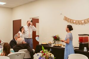 People gather at an indoor celebration with decorative banners and floral arrangements on tables in a community space.