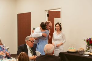 People gather at a celebration event with tables, flowers and cake in an indoor venue with brown doors.