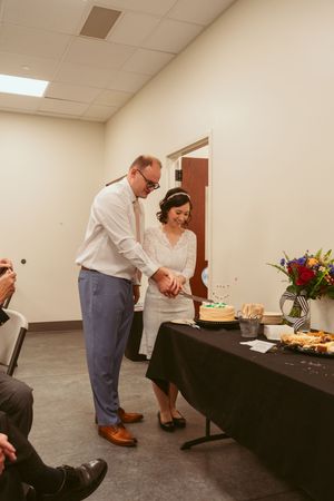 Two people select food items from a black tablecloth-covered buffet table in an indoor room with fluorescent lighting.