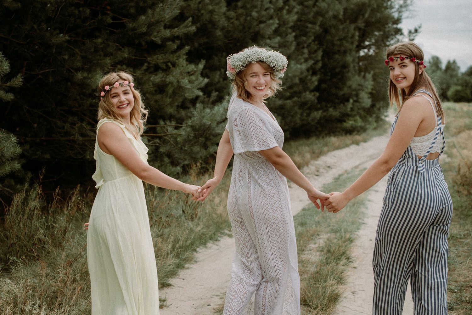 Friends in pastel dresses hold hands while walking along a countryside path at sunset.