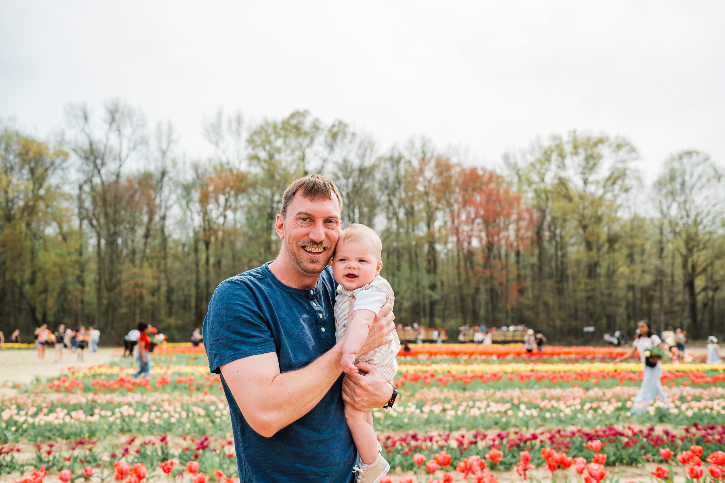Adult and baby pose together in front of colorful blooming tulip fields with autumn trees in the background.