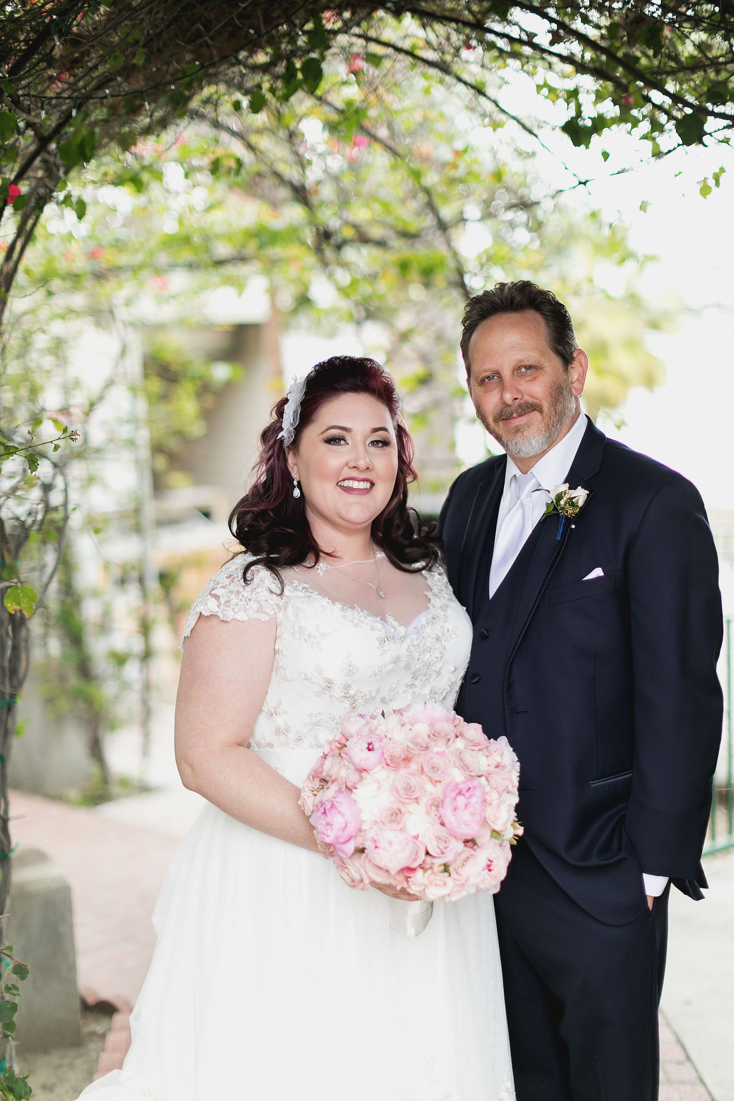 A wedding couple posing under tree branches with pink floral bouquet.