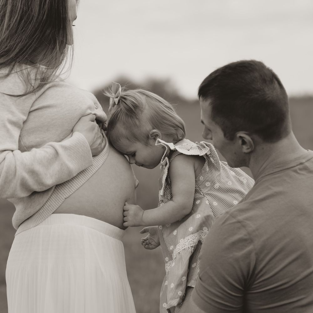 Black and white photo of parents holding their baby girl close to mothers pregnant belly outdoors.
