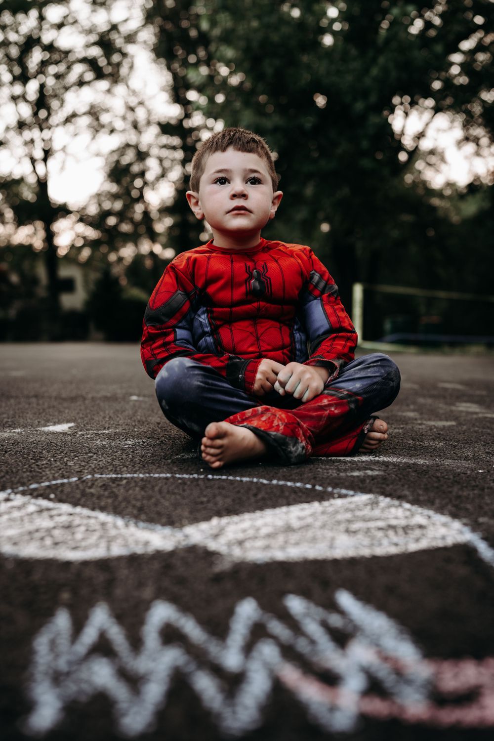 Child in Spider-Man costume sits cross-legged on pavement with chalk drawings.