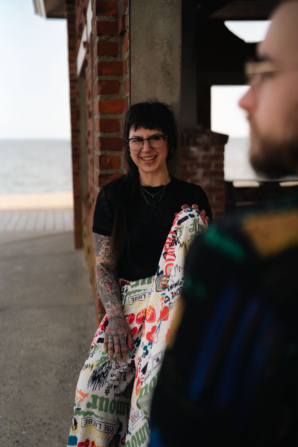 Two people stand together in a casual setting wearing denim and black clothing with tattoos visible. Engagement photoshoot engagement photog