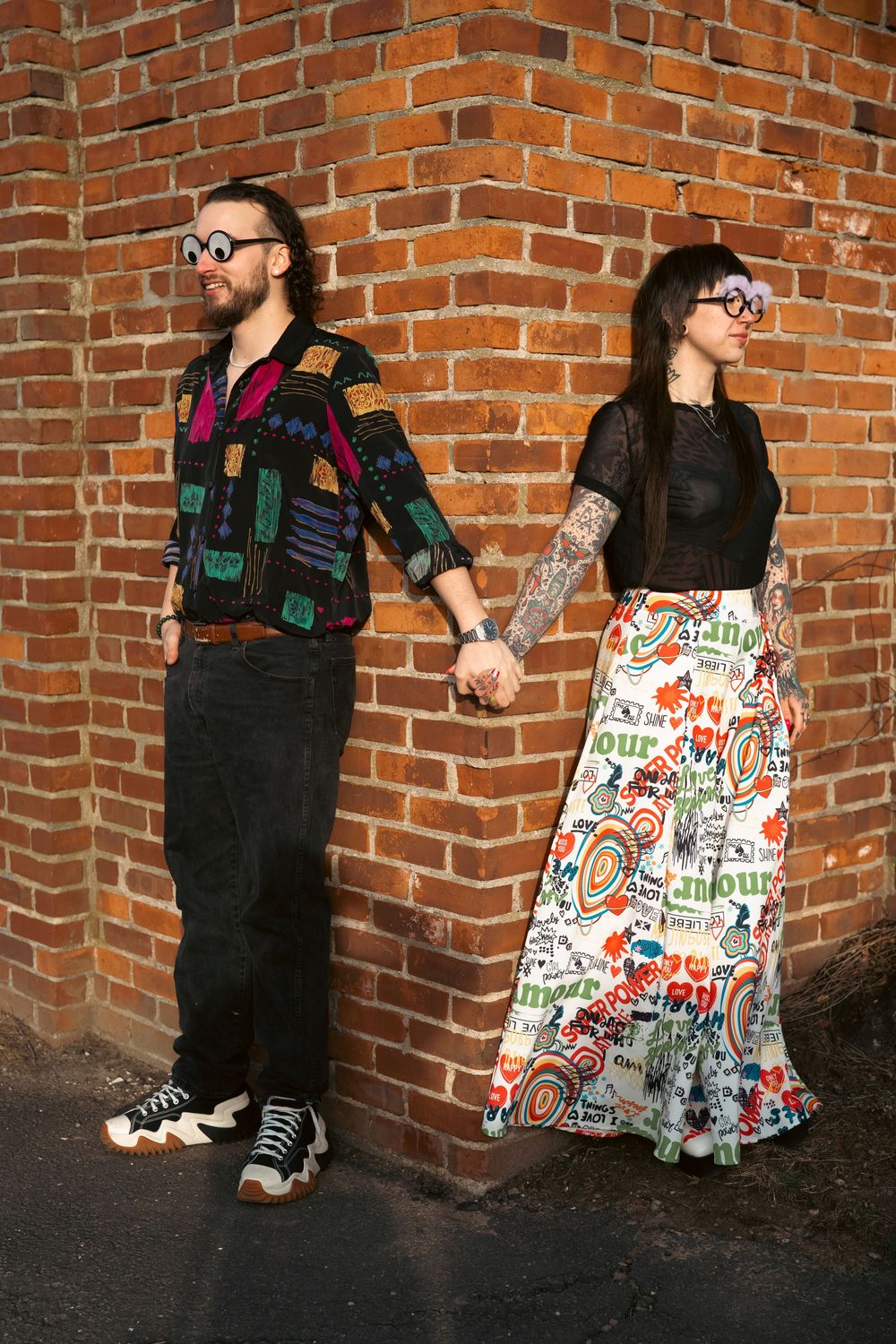 Two people pose playfully against a brick wall wearing contrasting black and patterned outfits. Engagement photoshoot engagement photography