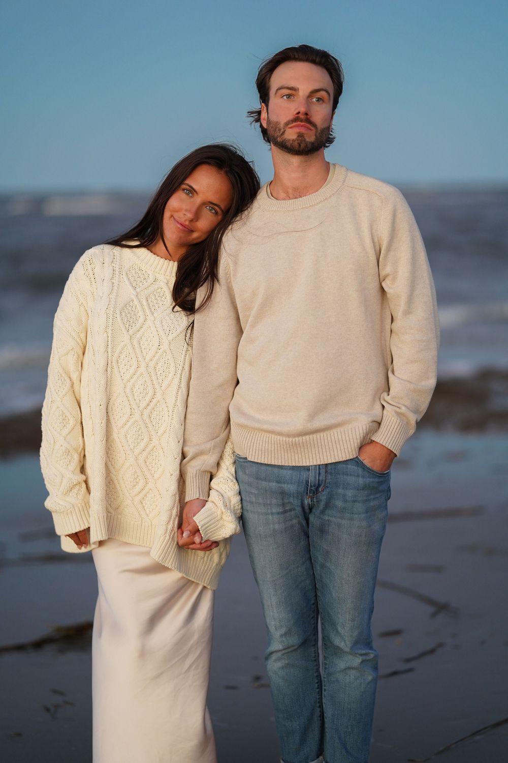 Couple in matching cream sweaters pose together on a beach at dusk.