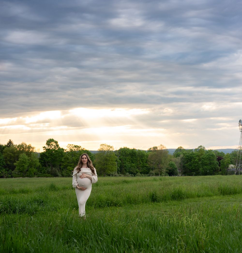 A pregnant woman in a white dress cradles her bump while standing in a green field at golden hour.
