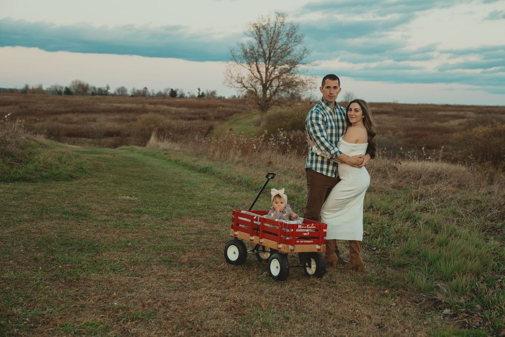 A couple poses for romantic outdoor photos in a rustic field with a red wagon on a cloudy autumn day.