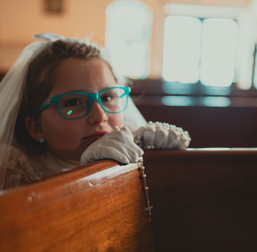 Young girl in First Communion veil and blue glasses holding a rosary while resting on a church pew.
