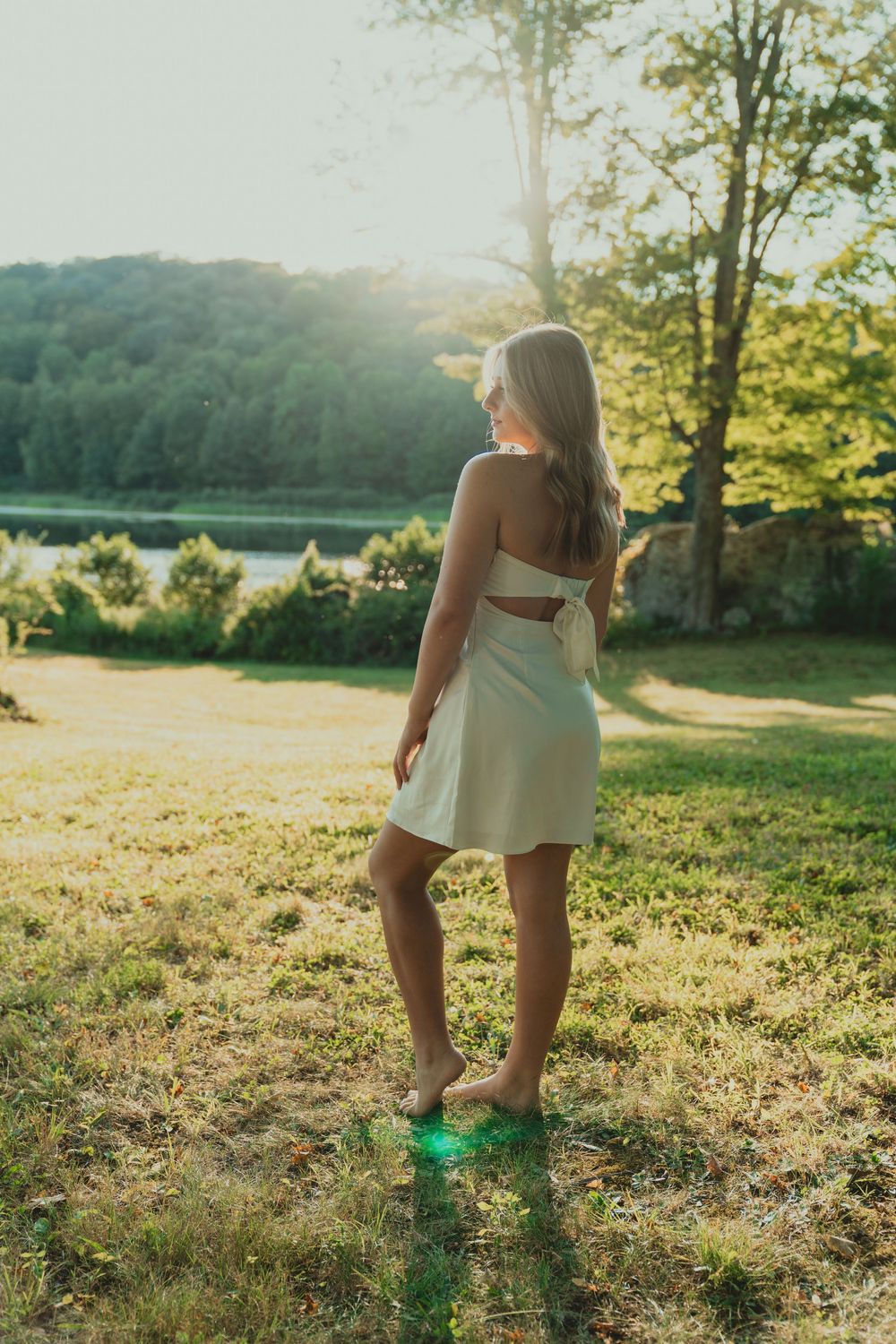 Figure in white dress and green boots stands in sunlit grassy field during golden hour.