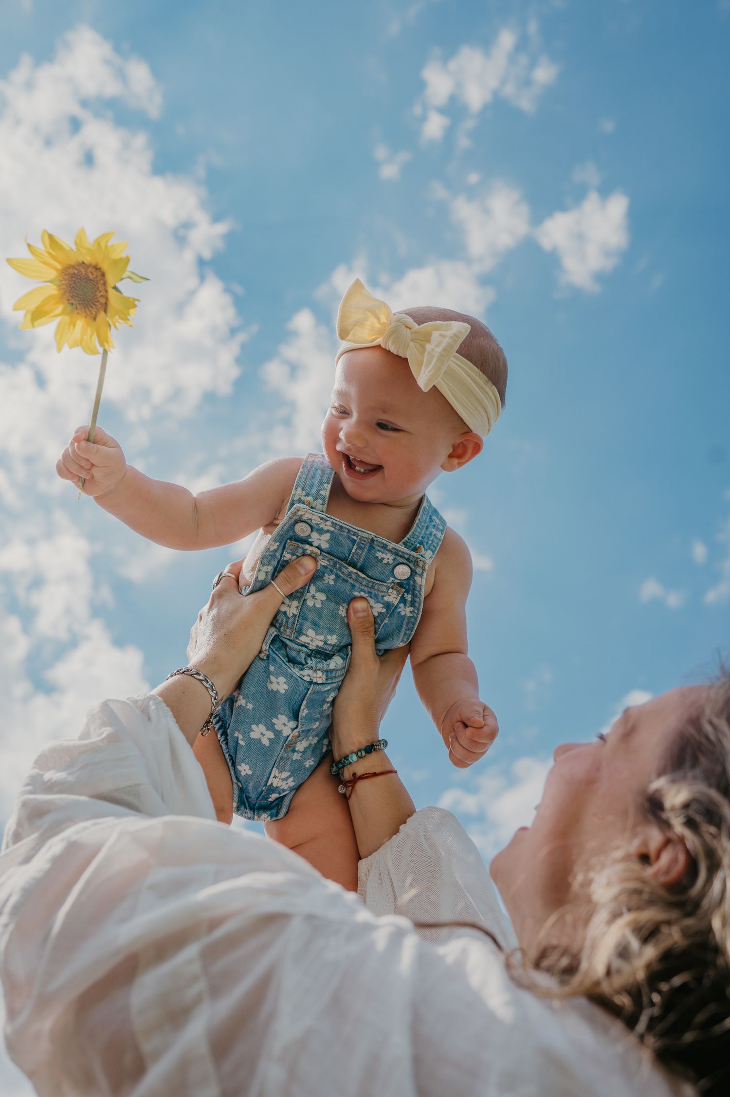 Joyful baby in denim overalls holding a sunflower being lifted up against a bright blue sky by a parent below.