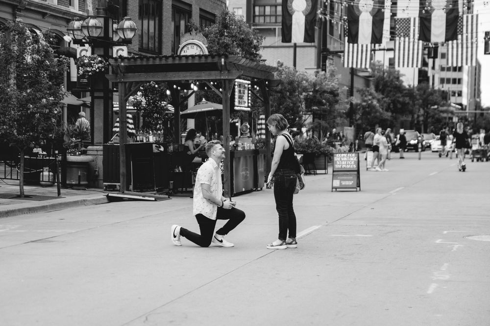 A black and white photo of a person proposing on a downtown street with American flags overhead.