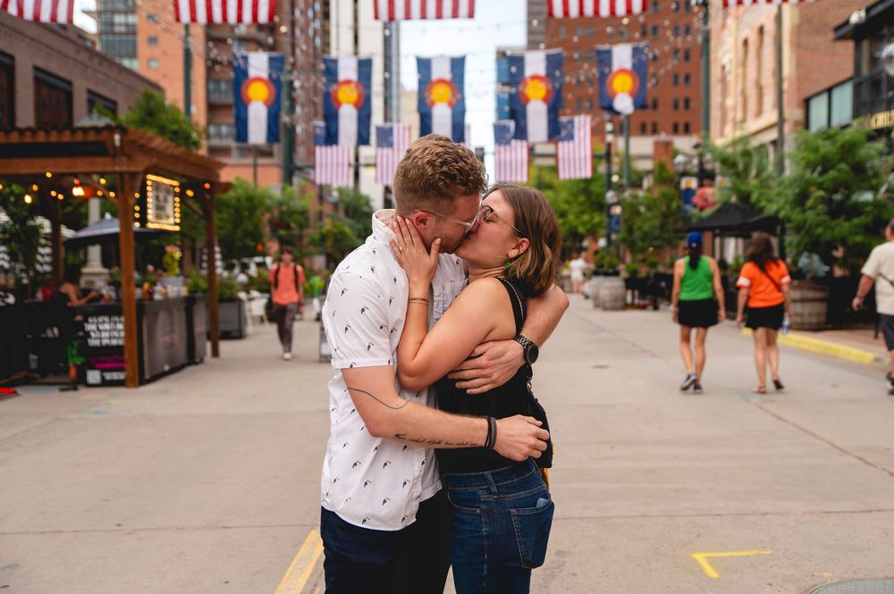 Young couple sharing an intimate embrace while standing on a city sidewalk near brick buildings and American flags.