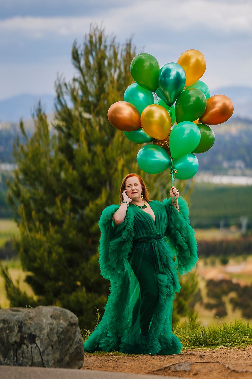 A person in a flowing emerald green dress poses with gold and green balloons against a scenic outdoor backdrop.
