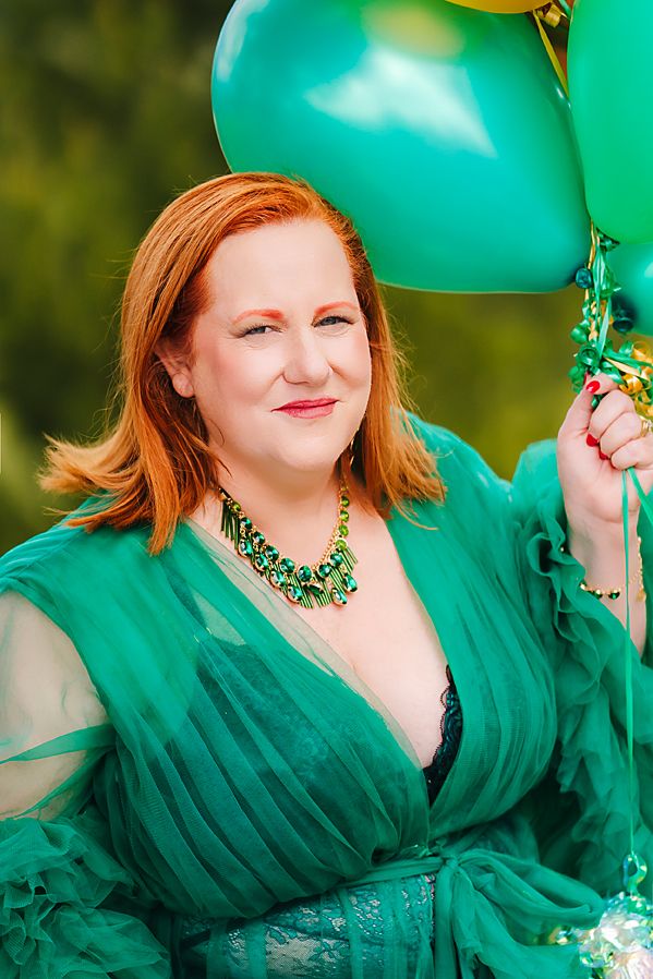 A model in a flowing emerald green dress poses gracefully with balloons against a blurred natural background.