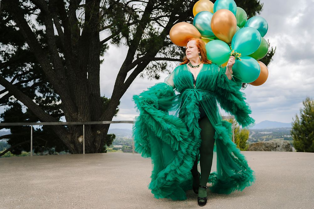 A person in a dramatic emerald green tulle gown poses with matching balloons against a cloudy sky.