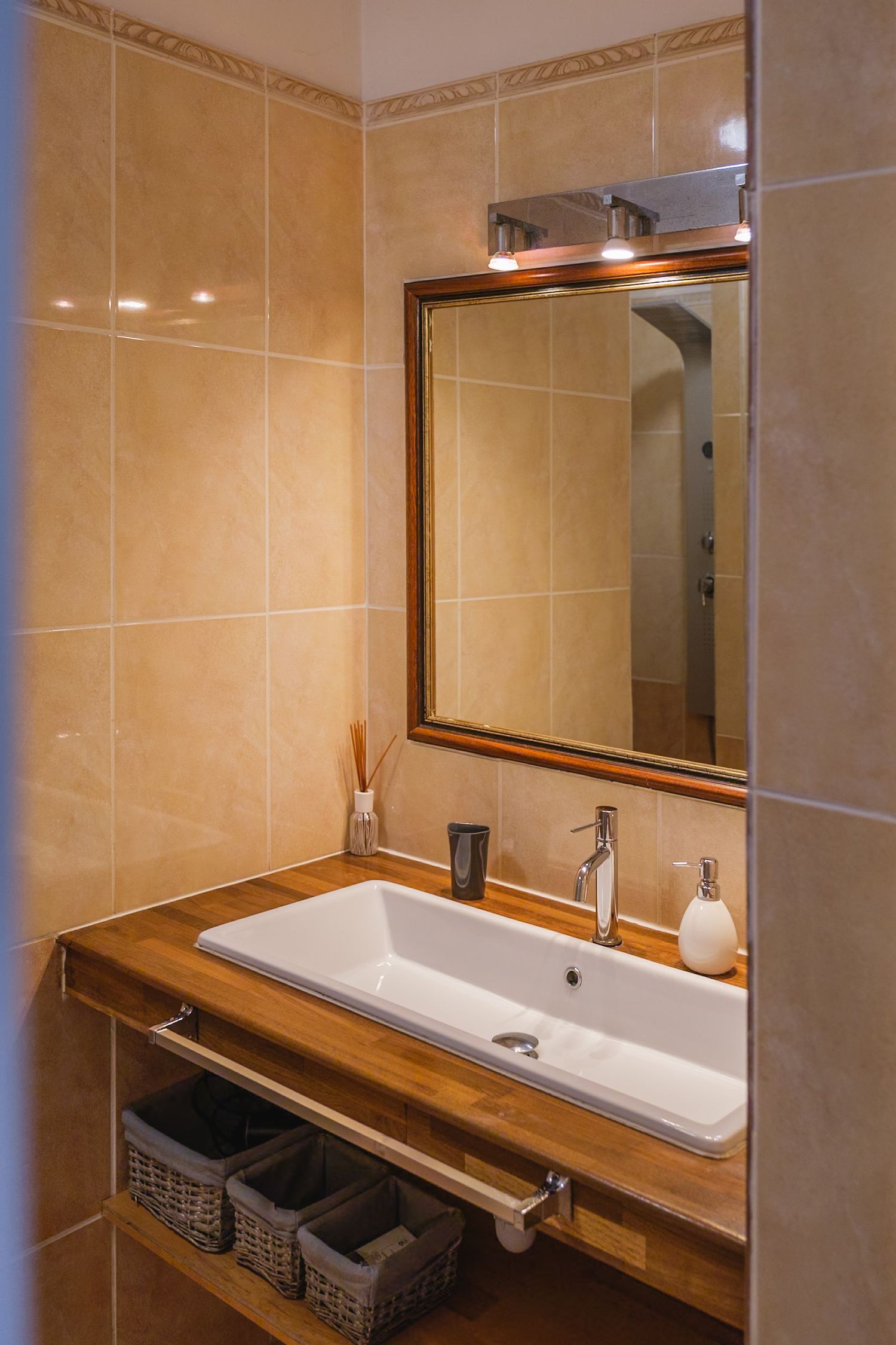 Modern bathroom vanity with wooden countertop, white sink, and framed mirror mounted on beige tiled walls with overhead lighting.
