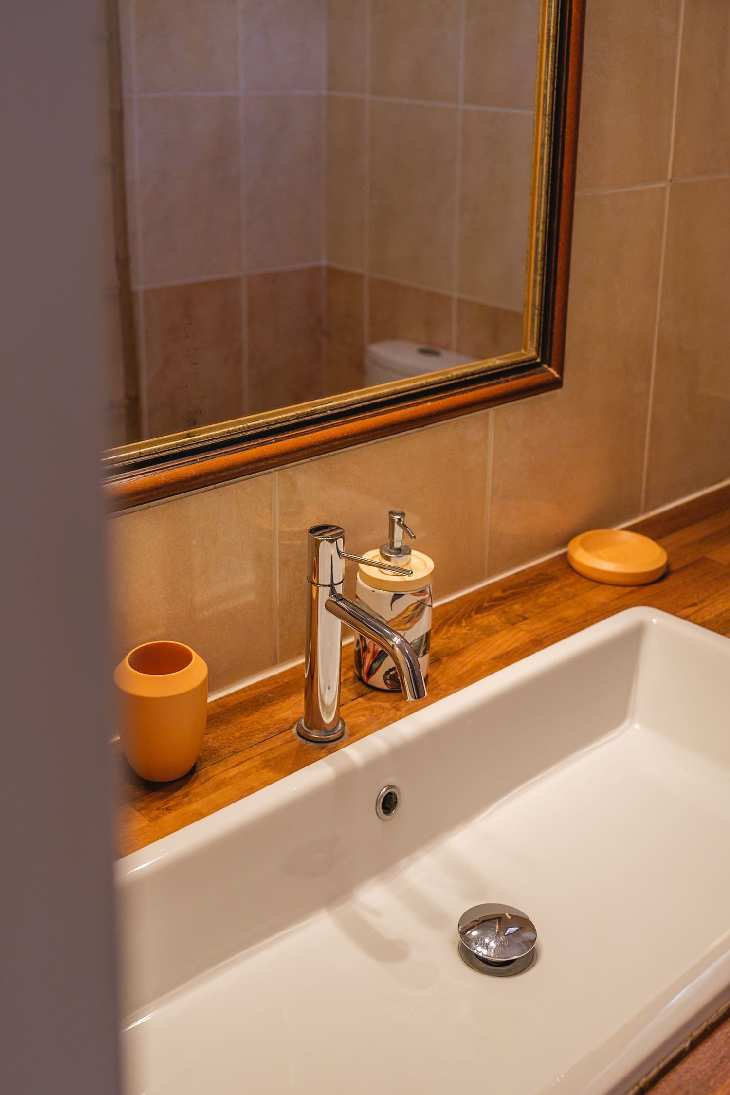 Modern bathroom interior with white ceramic sink, chrome faucet, wooden countertop and framed mirror on beige tiled wall.