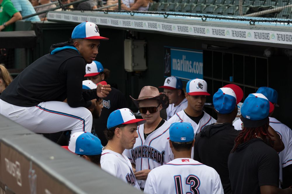 Baseball players in white uniforms sit in the dugout during a minor league game.