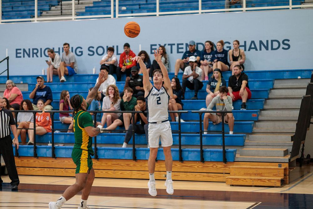 Basketball player takes jump shot during game with crowd watching in background.