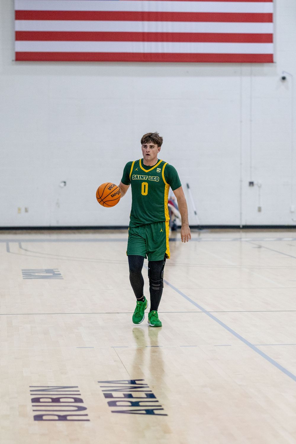 Basketball player in green uniform dribbles ball on court with American flag on wall.