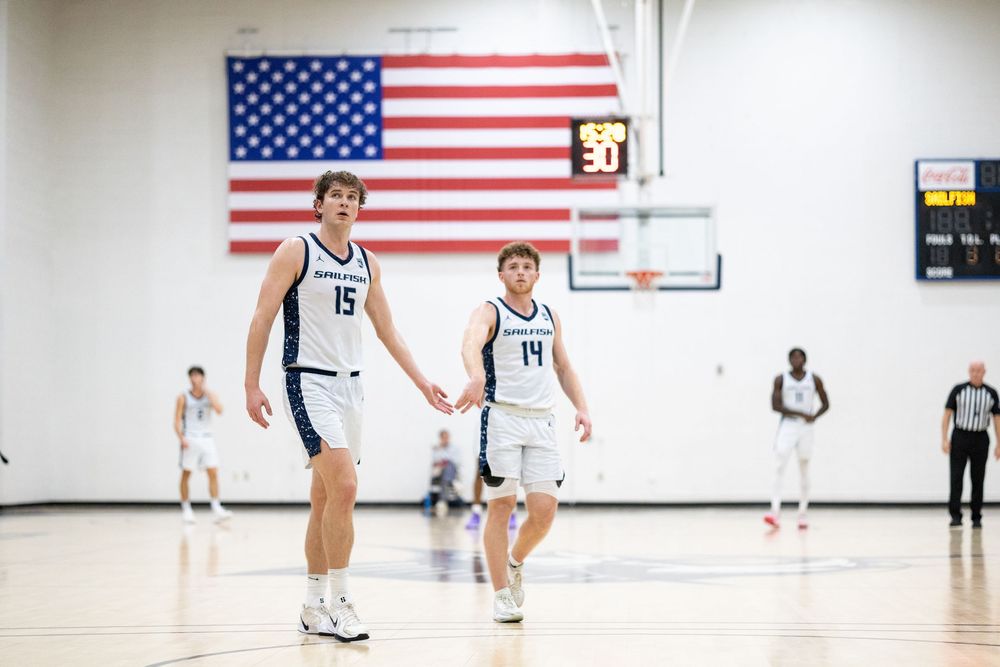 Two basketball players in white uniforms stand on court with American flag displayed.