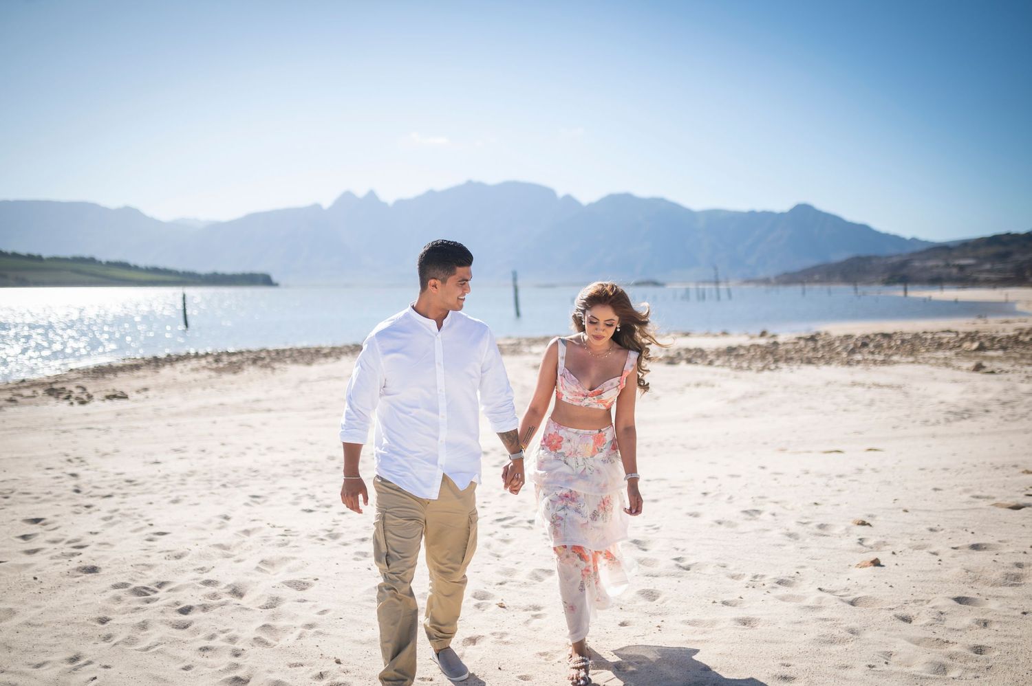 A couple walks hand in hand on a sandy beach with mountains and sparkling water in the background on a sunny day.