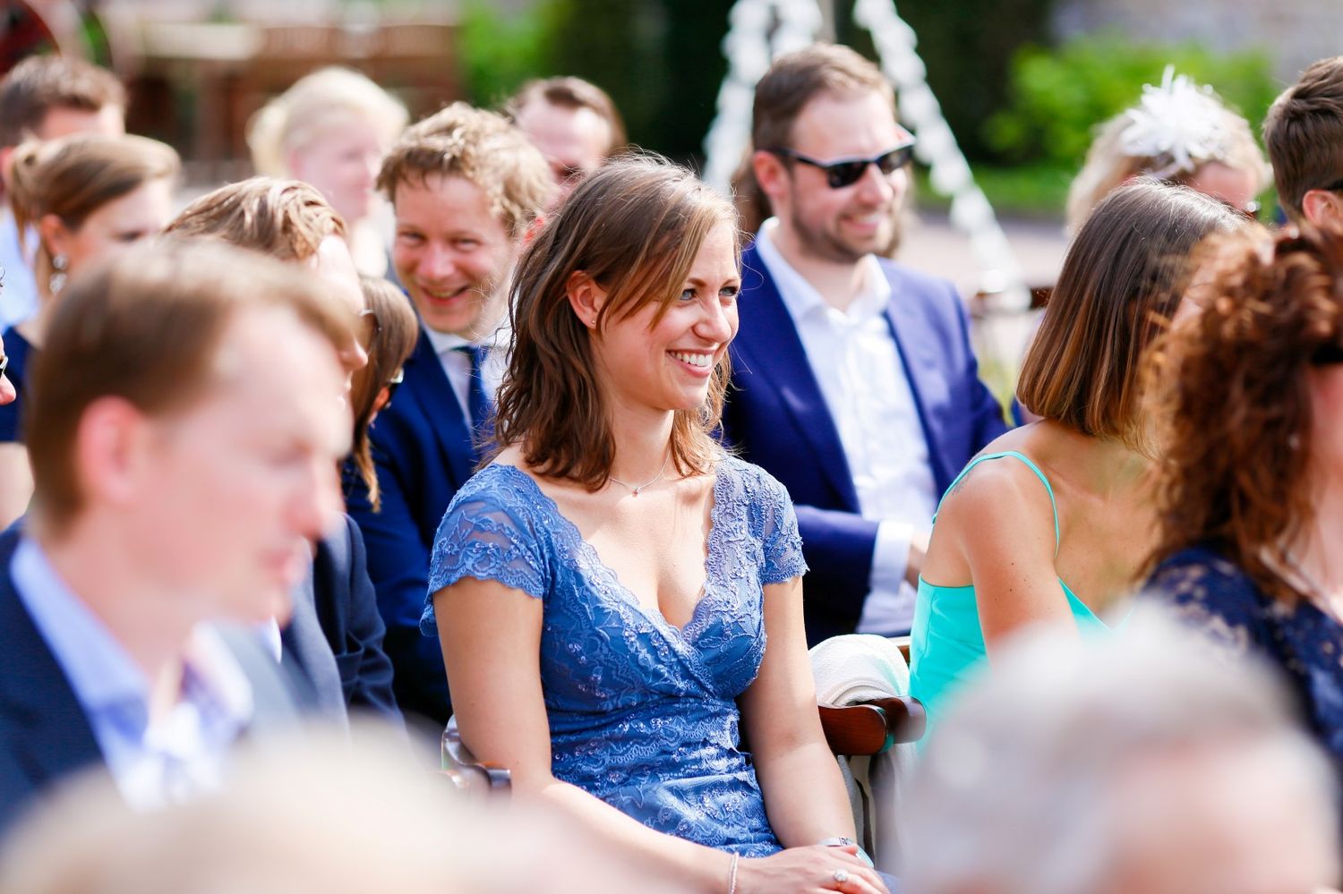 A lively outdoor gathering with people wearing formal attire including a sparkly blue dress in the crowd during a sunny day.