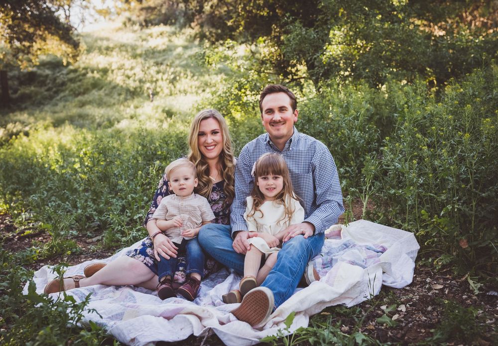 Family relaxes together on white blanket during golden hour outdoor photoshoot in wooded setting.