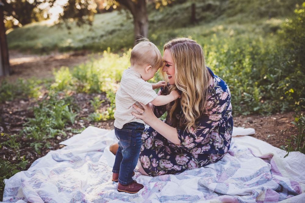 A parent and child sit together on a white lacy blanket outdoors surrounded by greenery and flowers.